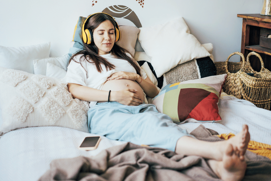 Pregnant woman lying on a bed with headphones, listening to a birth preparation class—taking in prenatal education through audio learning at home.