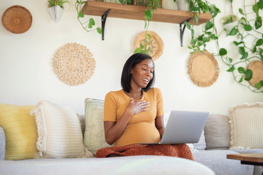 Pregnant woman in yellow top sits on a couch with laptop, smiling while accessing online content from home. Her cozy space features plants and woven wall decor, illustrating comfortable virtual learning during pregnancy.RetryClaude can make mistakes. Plea