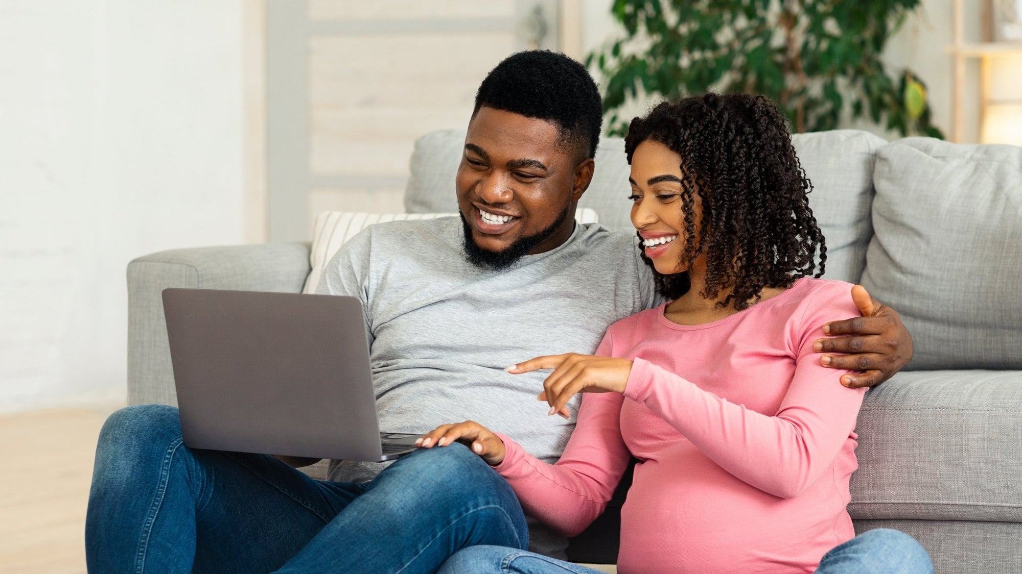 Expectant couple sitting on the floor, smiling and learning together on a laptop in a cozy home environment