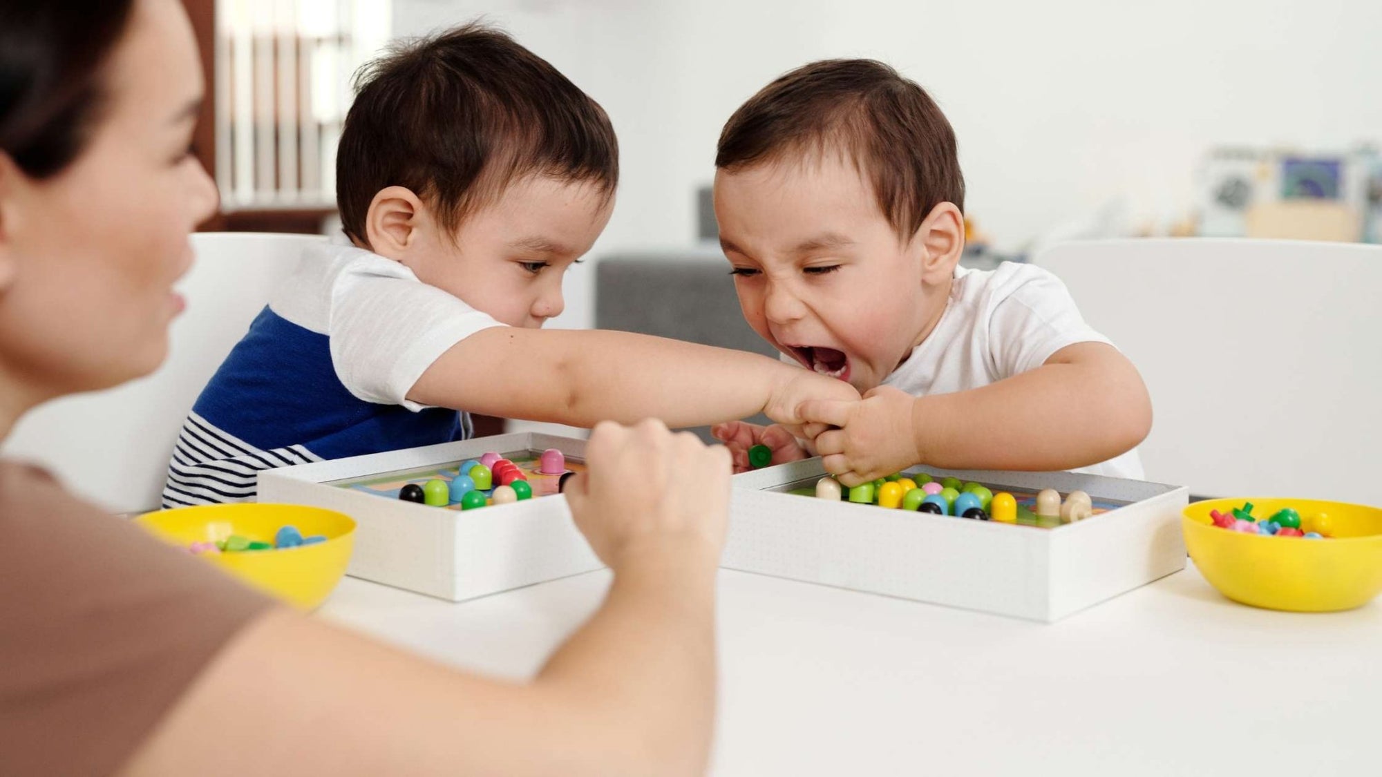 Two young children reaching into the same tray of colorful beads during a sensory activity, supervised by an adult