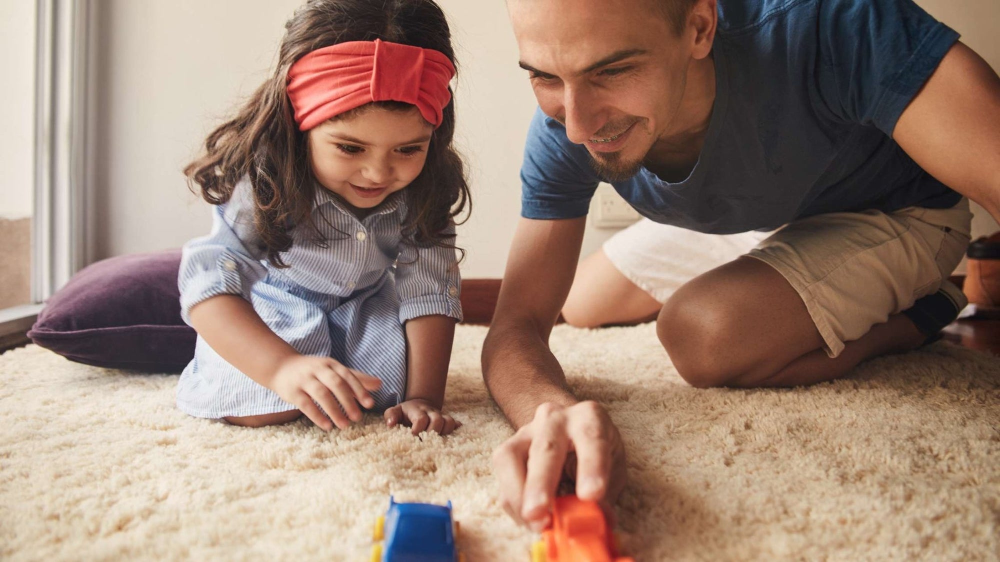 Father and young daughter smiling as they play together with toy cars on a soft carpet, showing engaged, screen-free playtime at home.