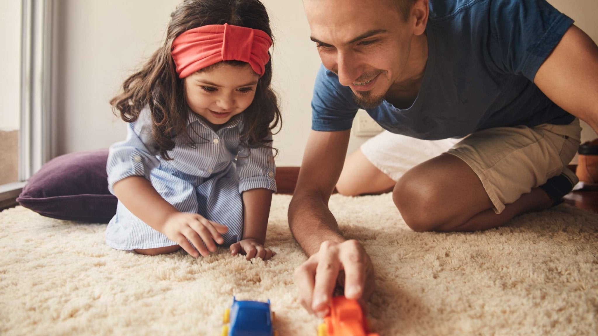 Father and young daughter smiling as they play together with toy cars on a soft carpet, showing engaged, screen-free playtime at home.